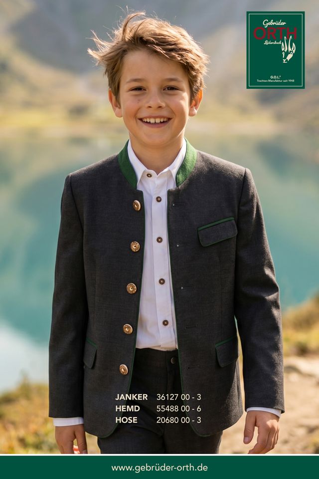 Junge in grauer Janker-Jacke mit grünem Hemd und schwarzer Hose, lächelnd in Berglandschaft.