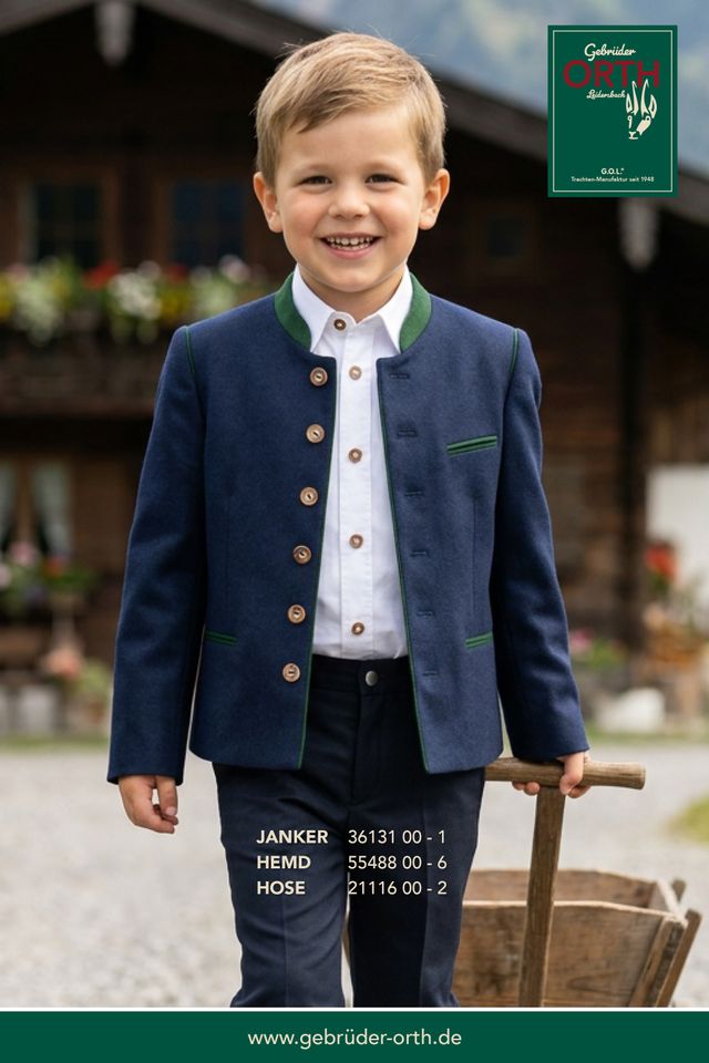 Junge in blauer Janker-Hose mit grünem Revers lachend vor Holzhaus.
