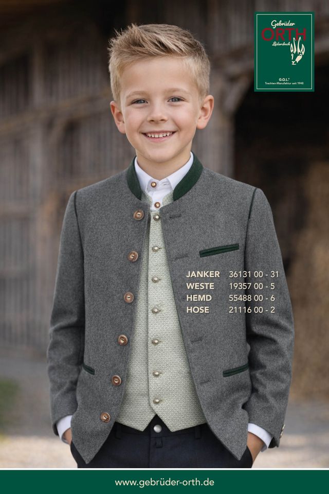 Junge in traditioneller Lederhose mit Weste und Jacke, lächelnd vor Holzhaus.