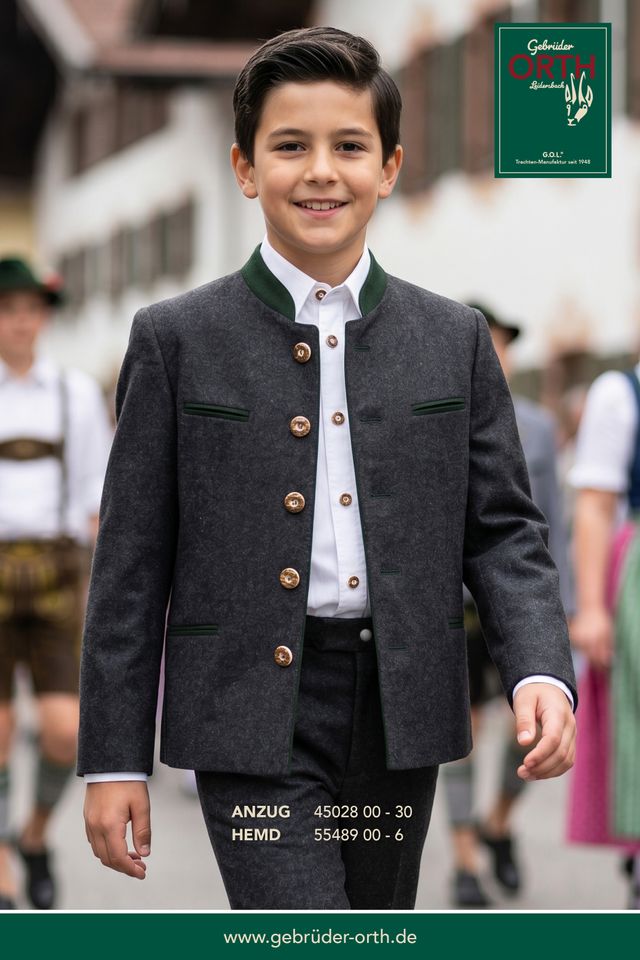 Junge in traditionellem Lederhosen-Jackett mit Weste und Hemd, lächelnd im Freien.