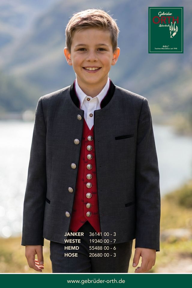Junge in grauer Janker-Weste mit roter Hose und Hemd, lächelt vor Berglandschaft.