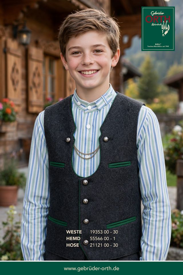 Junge in traditioneller Tracht mit Weste, Hemd und Hose, lächelnd vor alpiner Kulisse.